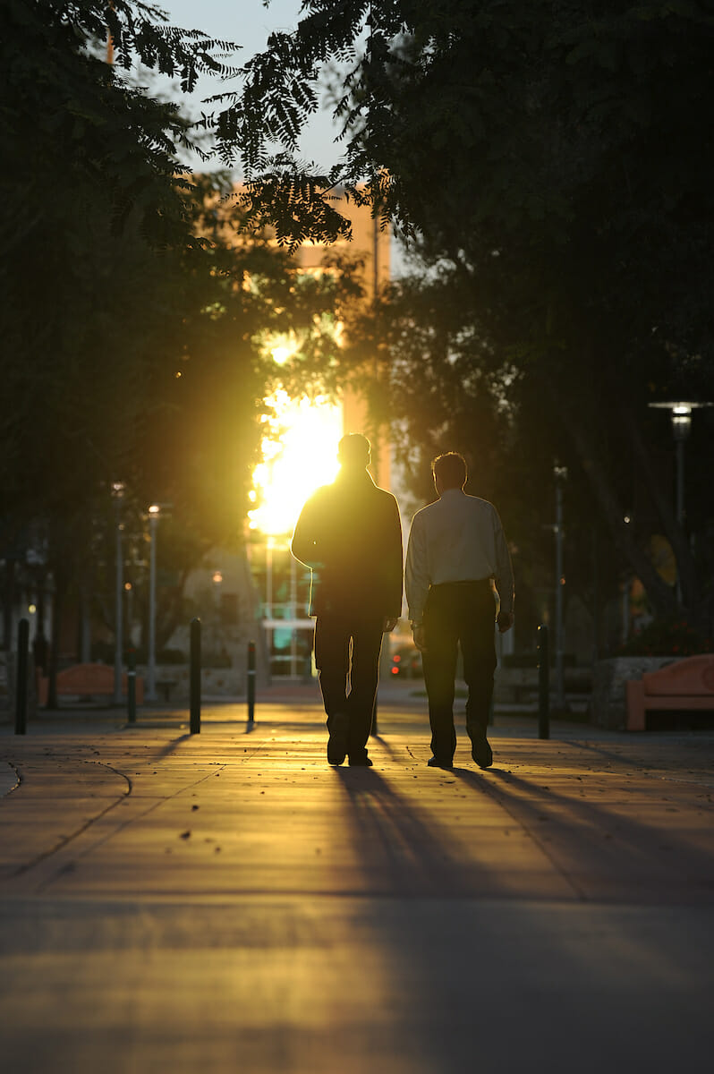 Two men walking down a sidewalk at sunset.