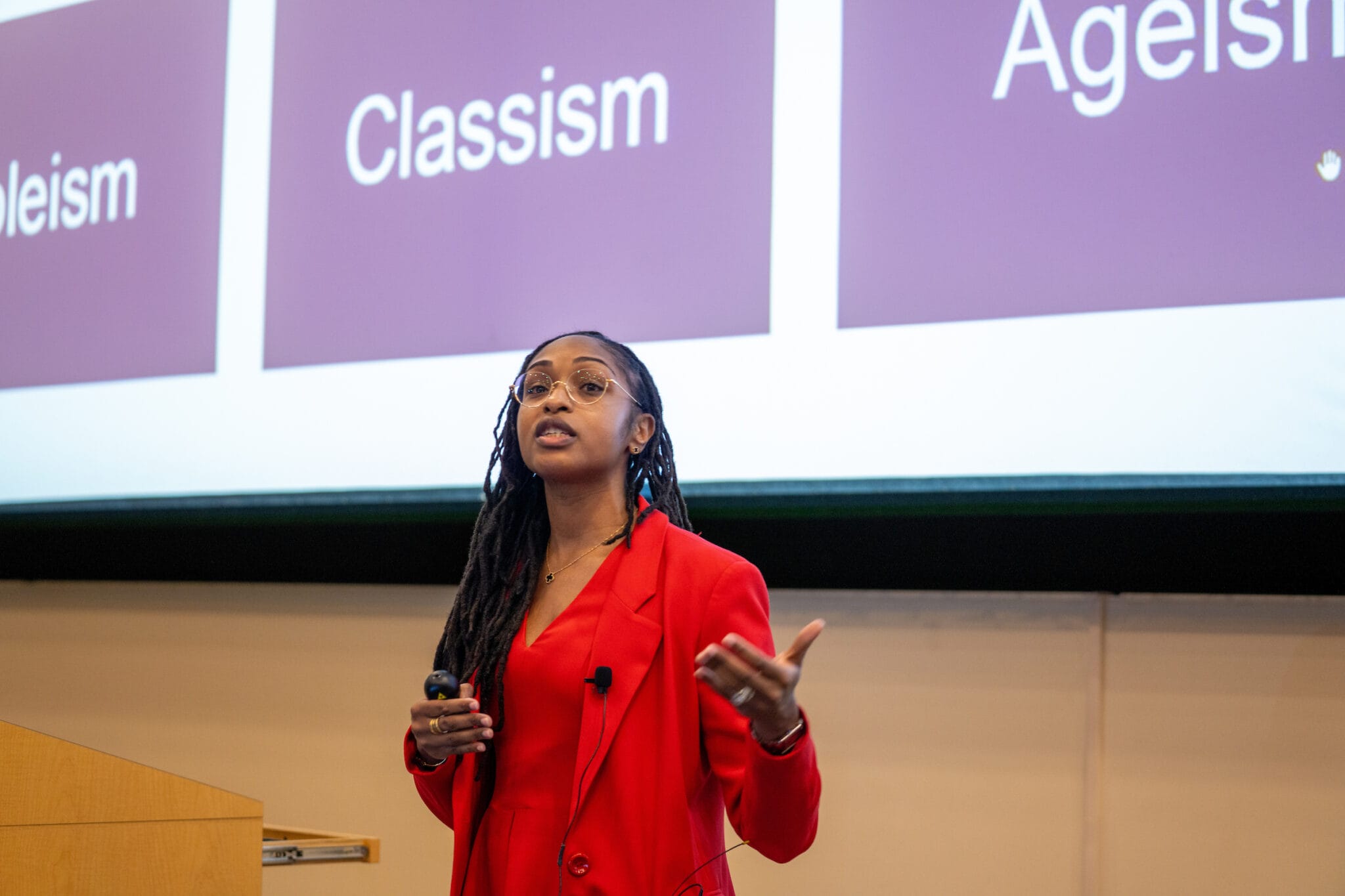 A woman in a red suit gives a presentation in front of a screen displaying the words "Ableism," "Classism," and "Ageism.