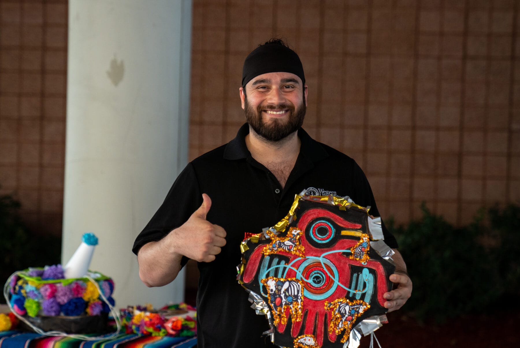 A man with a beard and a black headband gives a thumbs up while holding a decorated red mask. There are colorful items on a table behind him.