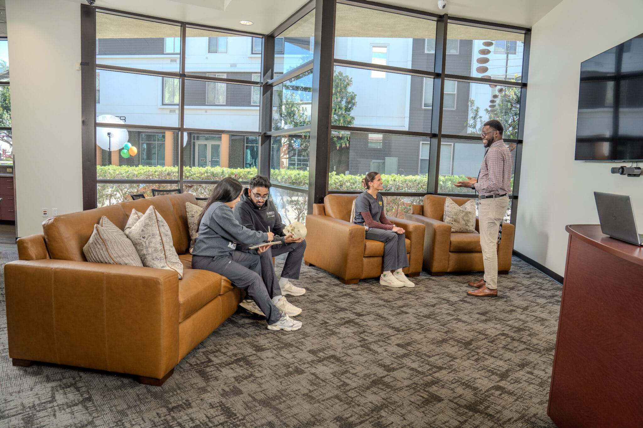 Four people are in a modern lounge area; three are seated on couches while one stands and speaks to the group. Large windows reveal an outdoor area with buildings and greenery.