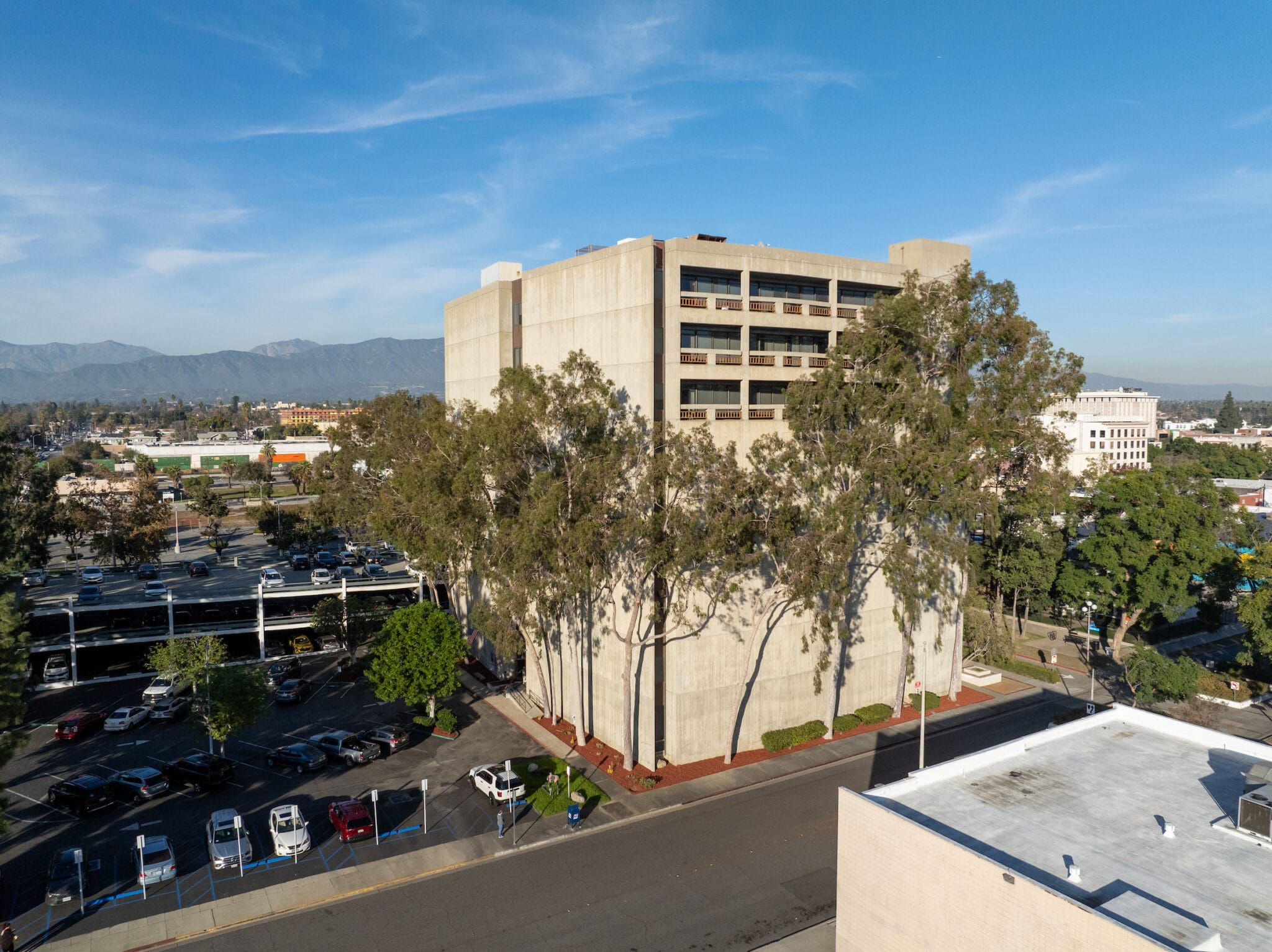 Aerial view of a multi-story concrete office building surrounded by tall trees, adjacent to a parking lot and city streets under a clear blue sky.