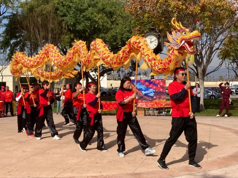 Participants perform a traditional dragon dance in a park, with several people holding a long, colorful dragon figure.