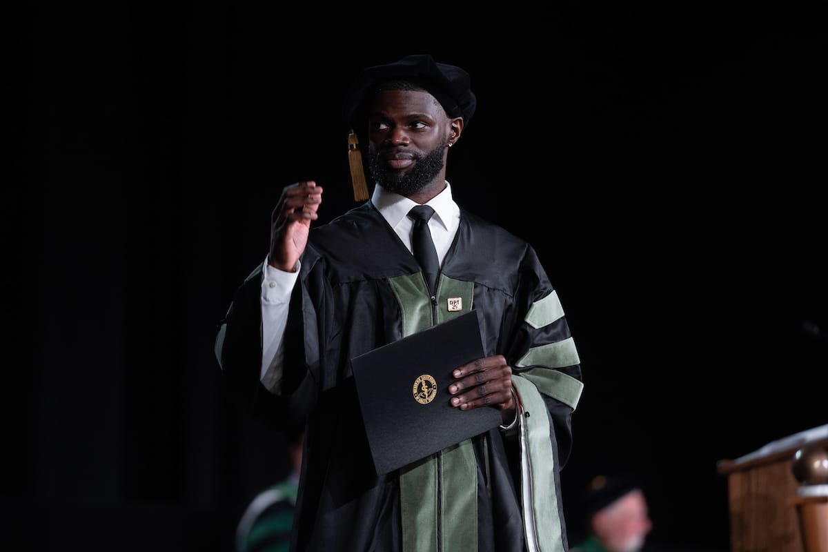 A person in graduation attire holds up their diploma on stage, dressed in black and green robes, with a black graduation cap with a tassel.