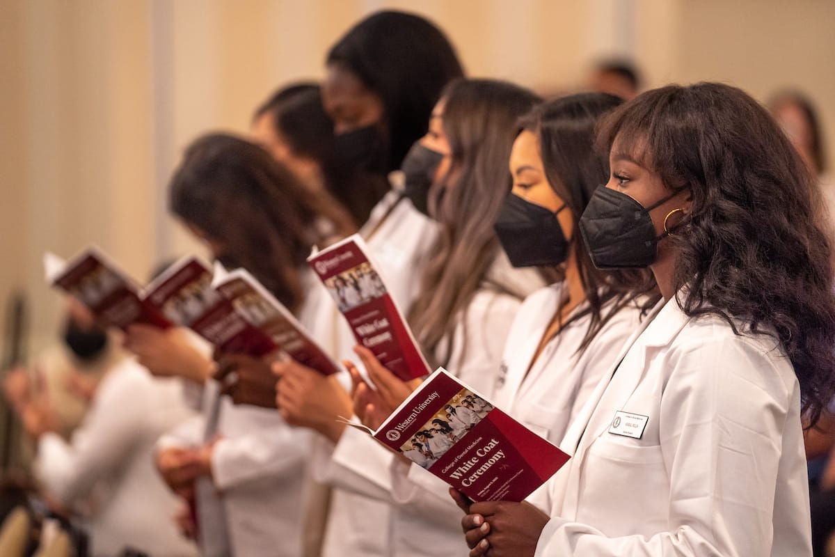 A group of people wearing white coats and black face masks stands in a line, holding and reading pamphlets.