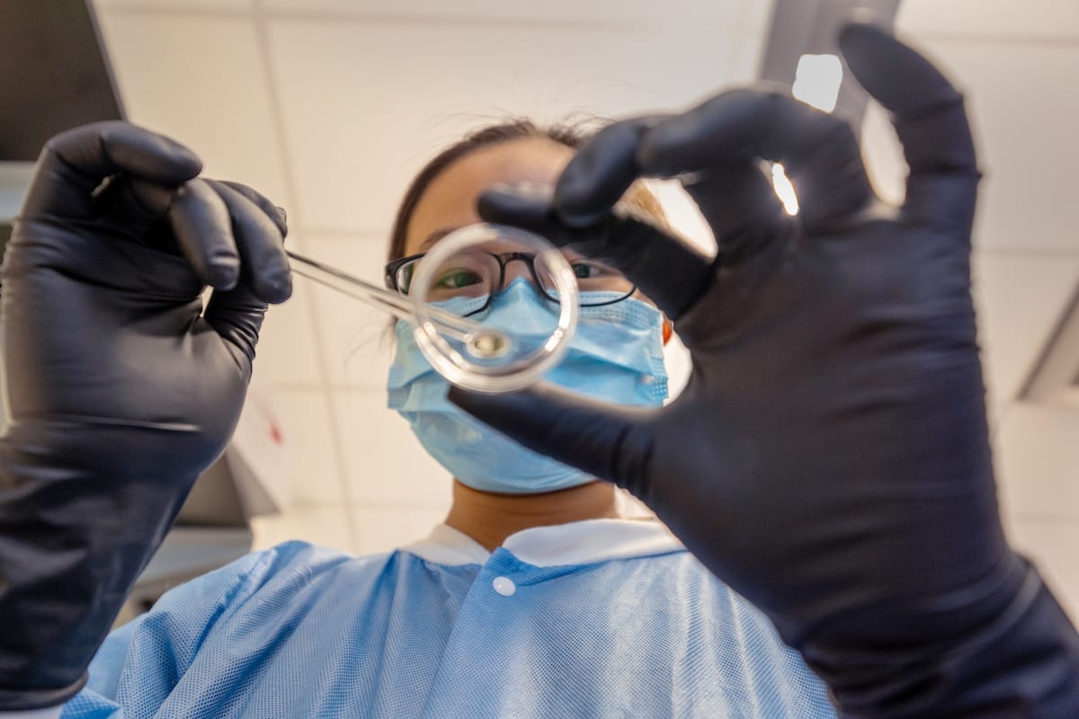 A person wearing a face mask, safety glasses, and black gloves holds a petri dish and tweezers in a laboratory setting.