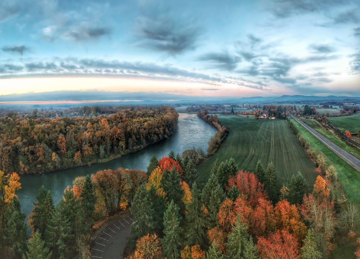 Aerial view of a winding river surrounded by autumn trees and farmland, with a road running alongside. The sky is partly cloudy, and the landscape features vivid fall colors.