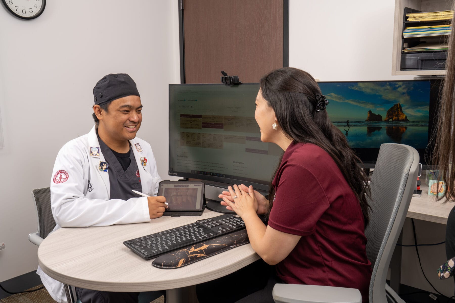 A healthcare professional shows a tablet to a seated woman during a consultation in a medical office with computers and paperwork visible.