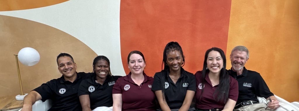Six people sit on a couch, smiling at the camera, in front of a wall with orange and beige abstract shapes.