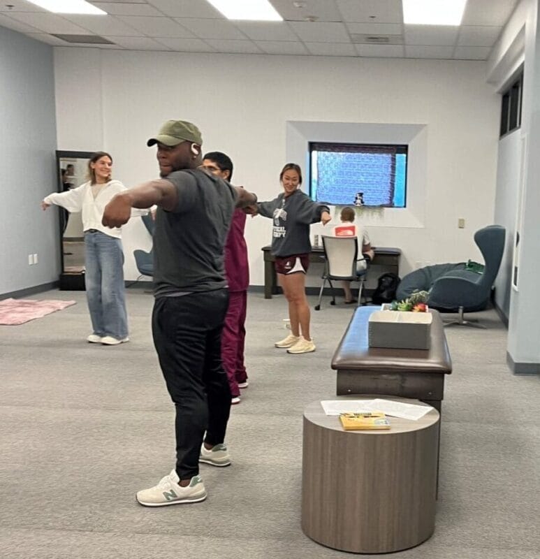 A group of people stands in a room with arms outstretched, participating in a group activity or exercise. Tables and chairs are visible in the background.