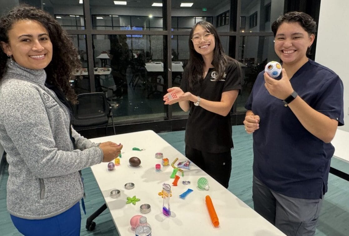 Three people stand around a table covered with sensory toys and objects, smiling at the camera in a brightly lit indoor setting with large windows.