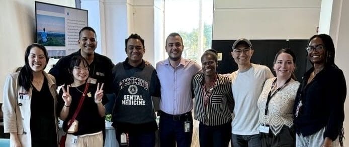 A group of nine people stand together smiling in an office setting with a TV, chairs, and large windows in the background.