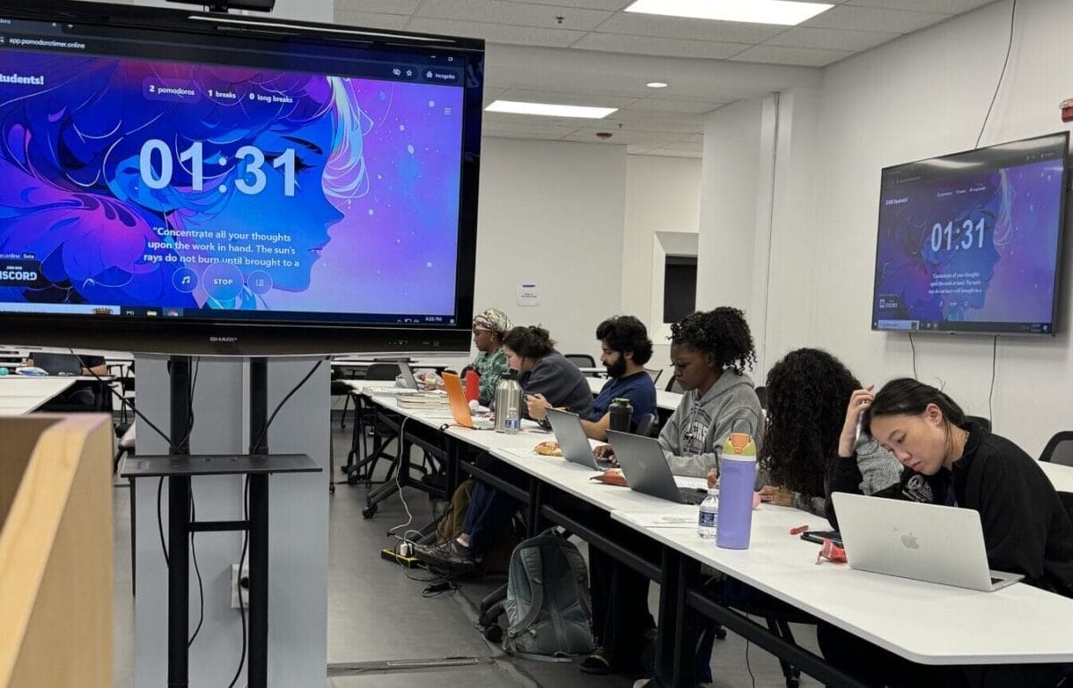Several people sit at a long table using laptops in a classroom. A large screen displays a timer and colorful graphics at the front of the room.