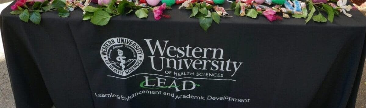 A table covered with a Western University of Health Sciences LEAD tablecloth displays colorful eggs, candies, a water bottle, a metal bucket, and informational materials outside.