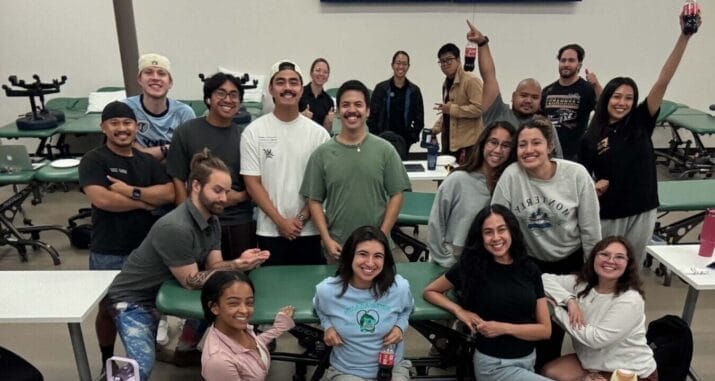 A group of people in a classroom pose for a selfie, with desks, chairs, and two monitors displaying a blue screen in the background.