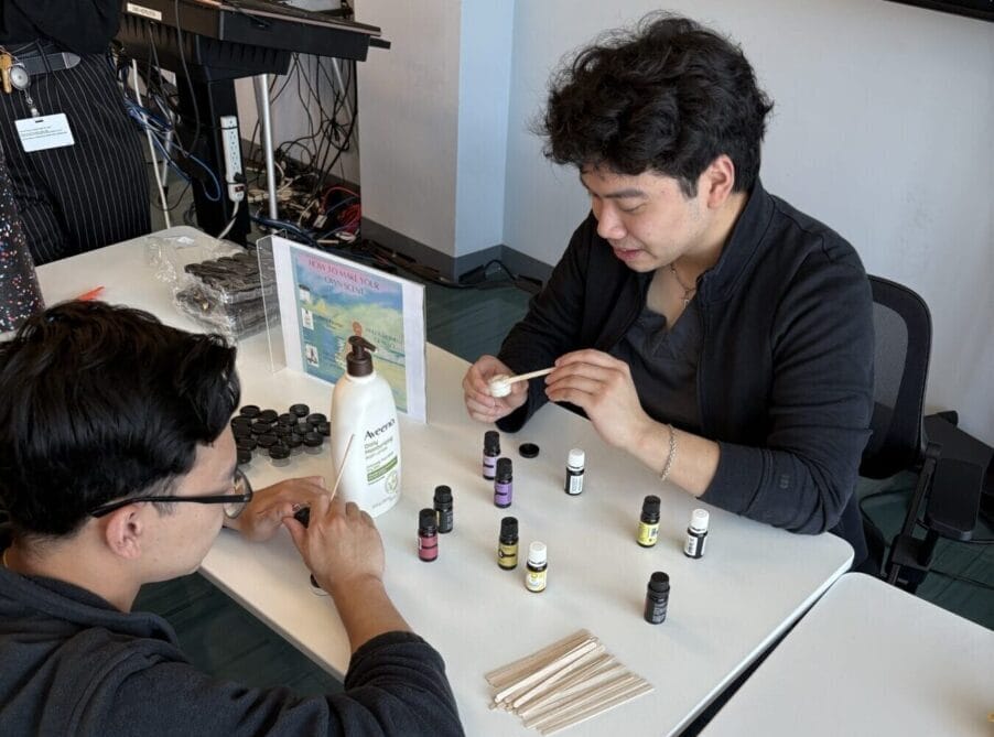 Two people sit at a table arranging essential oil bottles and wooden sticks, with other supplies and a bottle of lotion nearby.