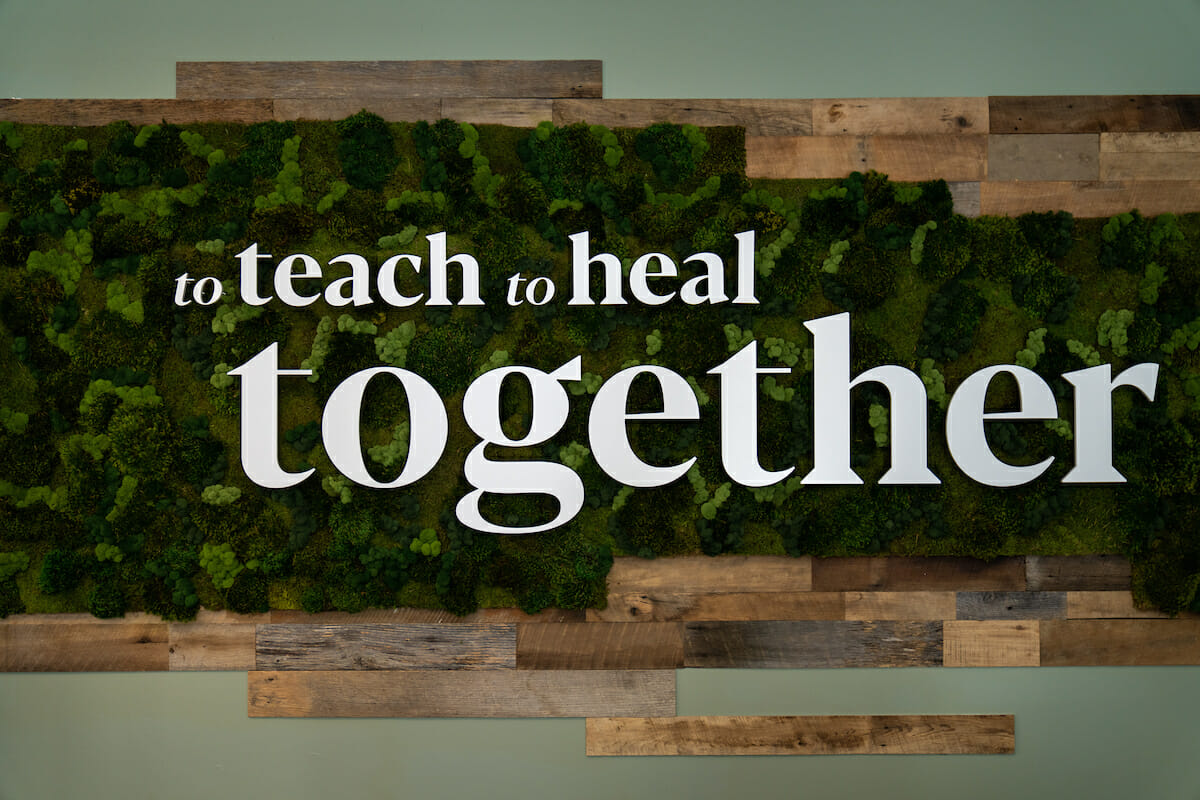 A sign with the phrases "to teach to heal together" is displayed on a green, leafy background with wooden accents.