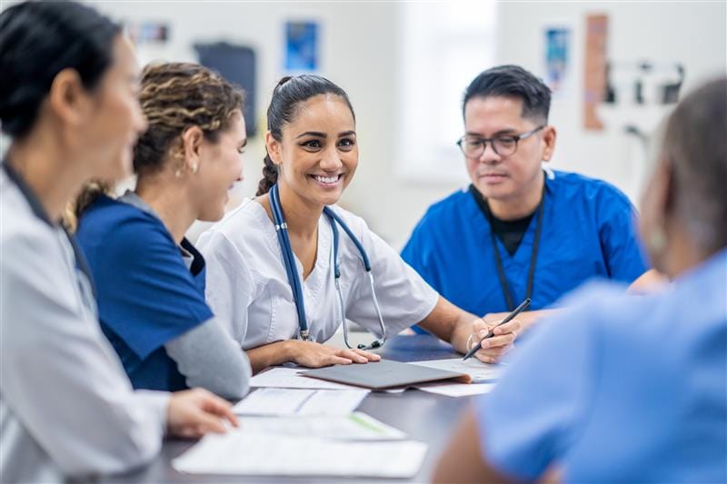 A group of healthcare professionals in scrubs sit around a table, talking and reviewing documents together in a bright room.
