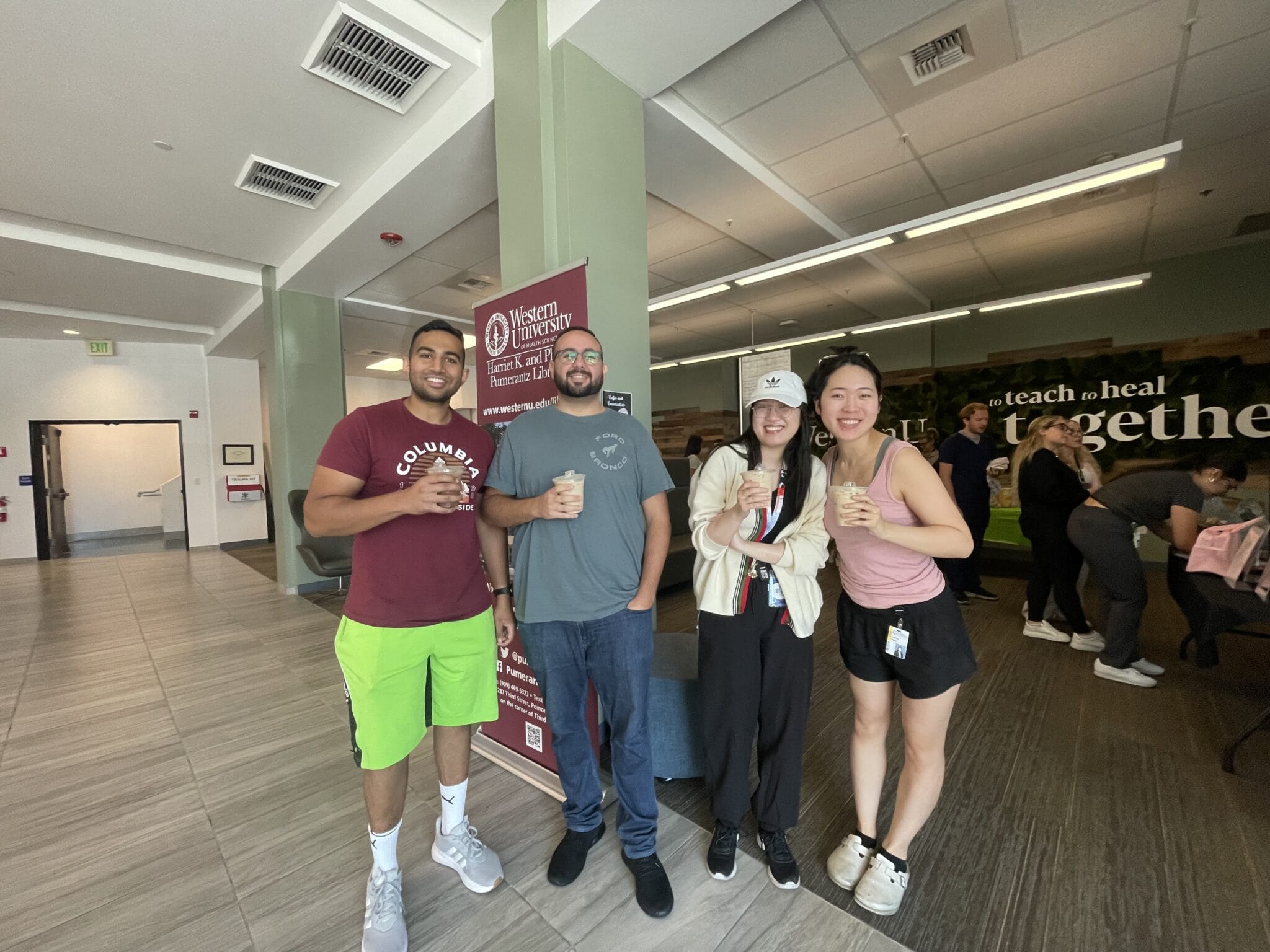 Four people stand together indoors, smiling and holding drinks, with a Western University banner and people in the background.