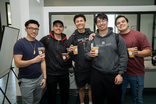 Five young men stand indoors, smiling and holding iced coffee drinks, dressed in casual clothing and hoodies.
