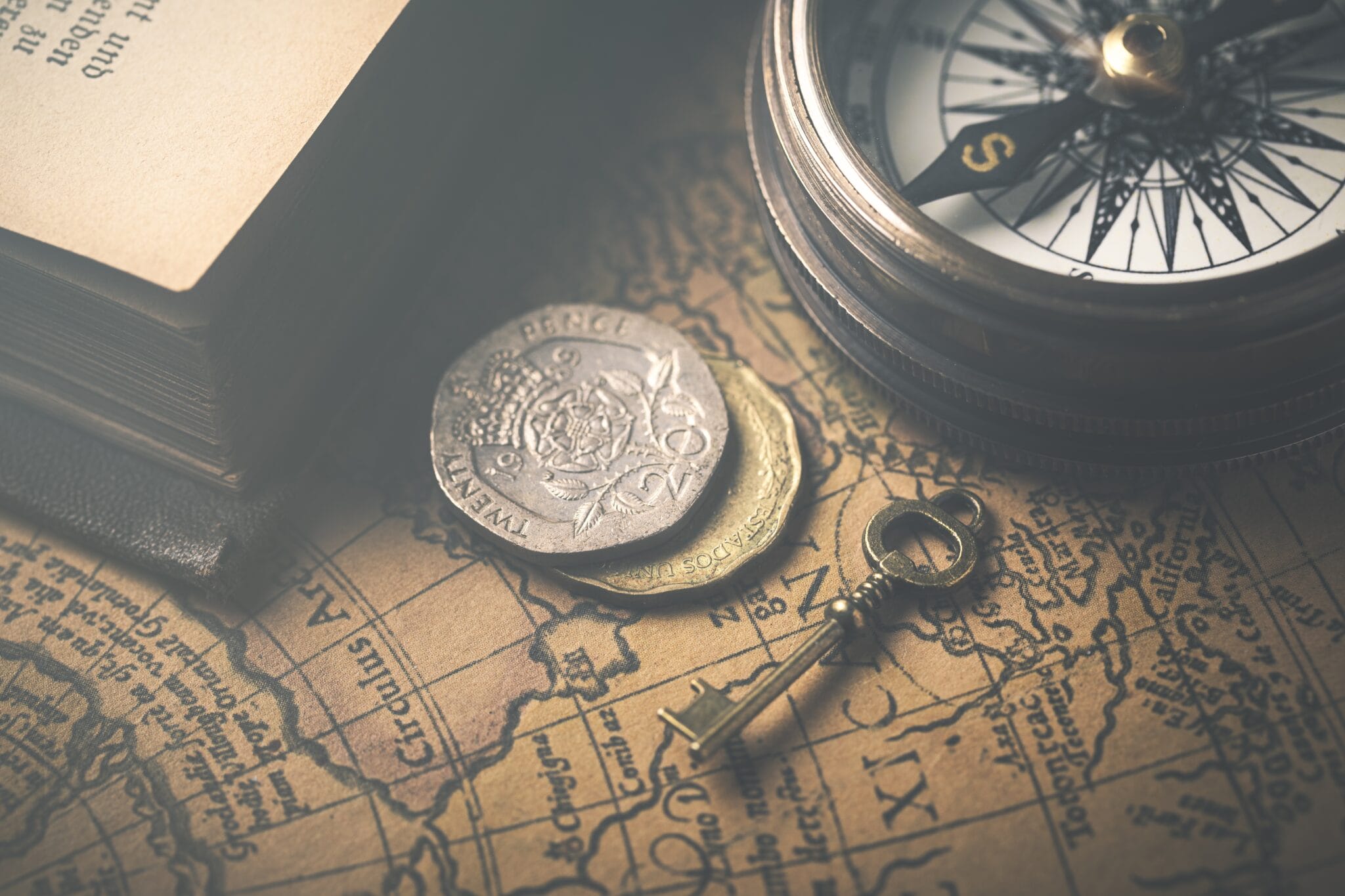 Old coins, a vintage key, and a compass rest on an antique map next to an open book.