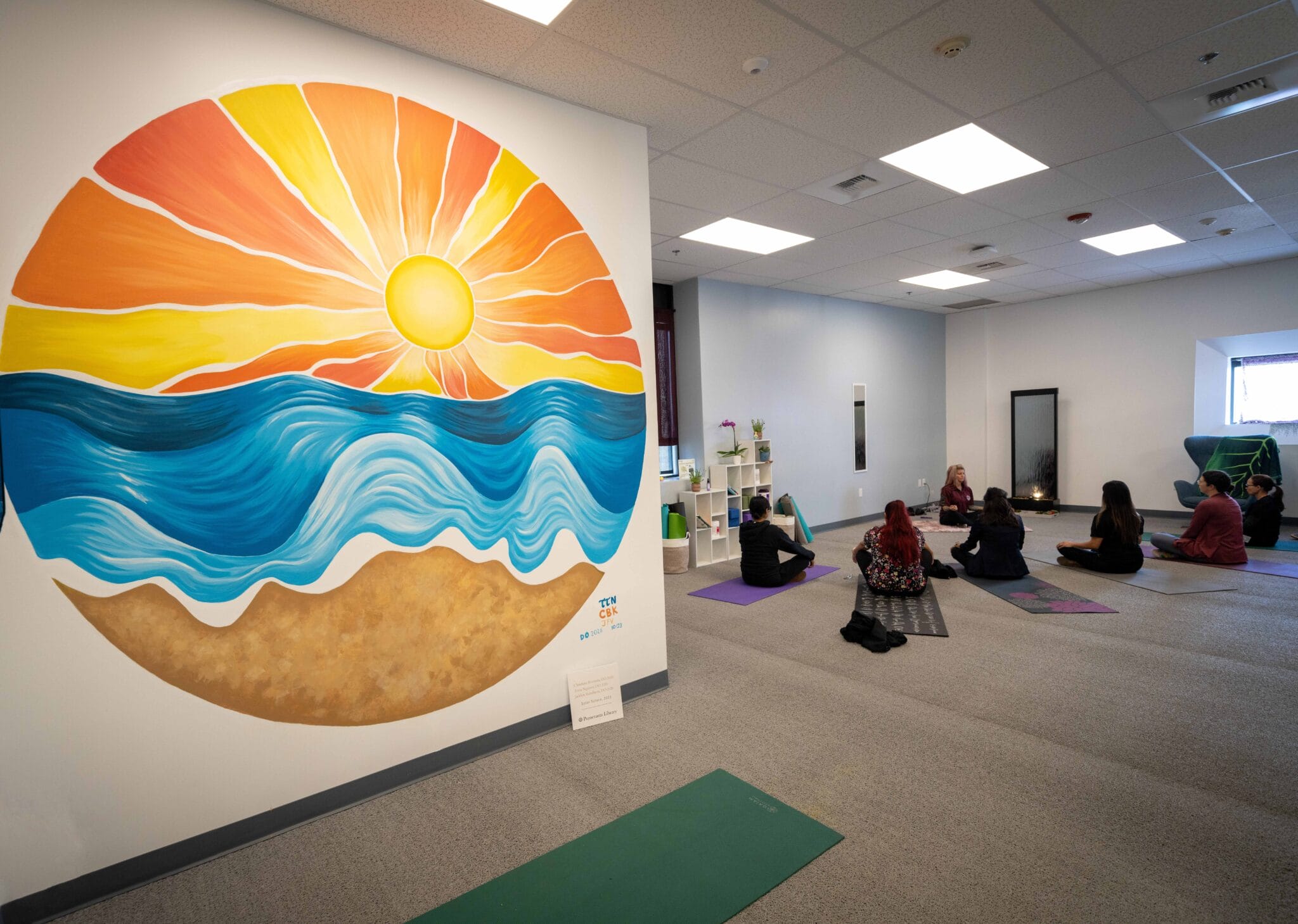 A group of people sit on yoga mats in a room with a large mural of a sun, ocean, and sand on the wall.