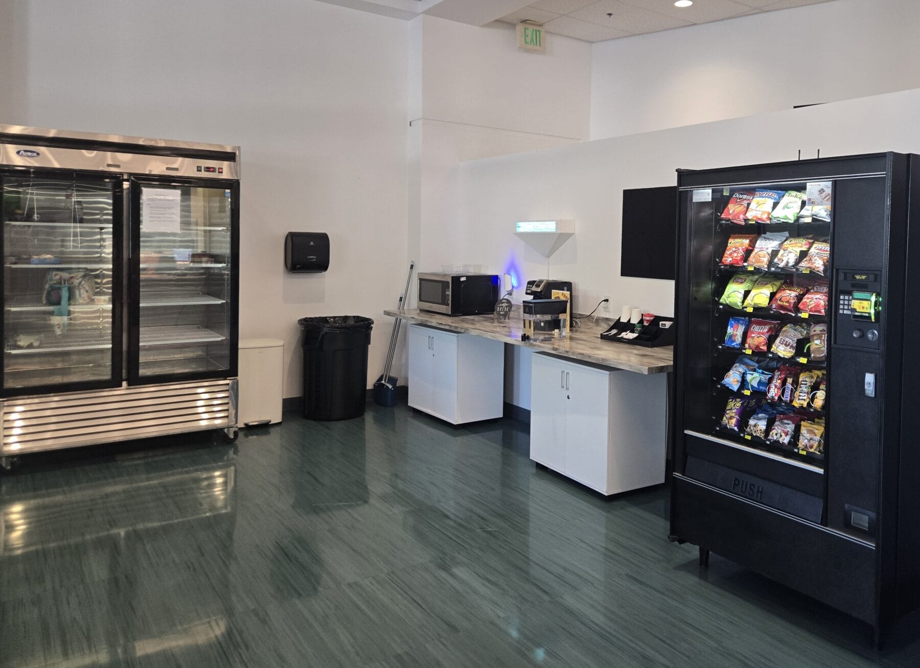 Break room with a refrigerator, a vending machine with snacks, a countertop with microwaves, sink, paper towel dispenser, and trash can on a shiny green floor.