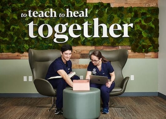 Two people in blue scrubs sit in chairs working on a tablet and notes in front of a wall with the text "to teach to heal together" in an office setting.