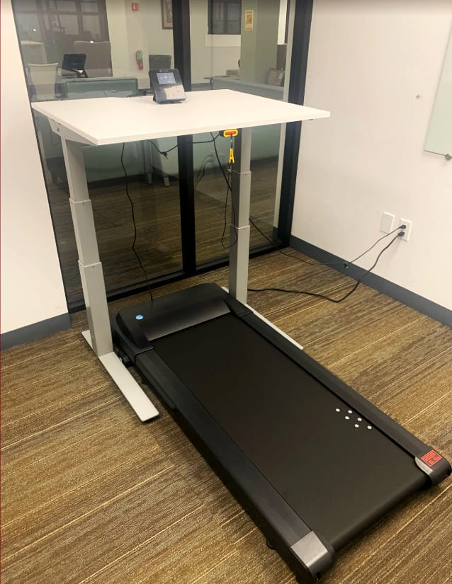 A treadmill desk setup in an office with a white height-adjustable desk and a black treadmill below it, against a window and adjacent to a power outlet.