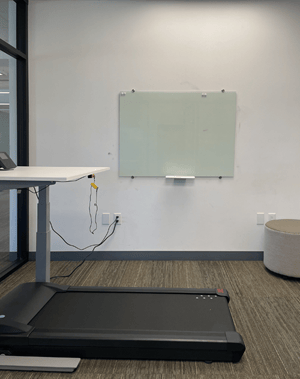 Office space featuring a standing desk with connected cables, a glass whiteboard on the wall, a treadmill in front of the desk, and a round ottoman in the corner.