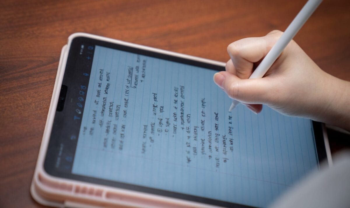 A person writes notes on a tablet using a stylus. The tablet displays lined digital paper with handwritten text in English.