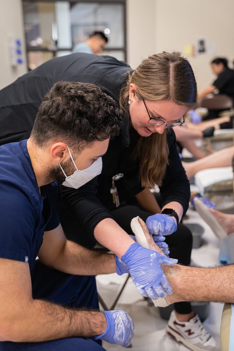 Two healthcare workers are applying a cast to a patient's leg in a clinical setting.