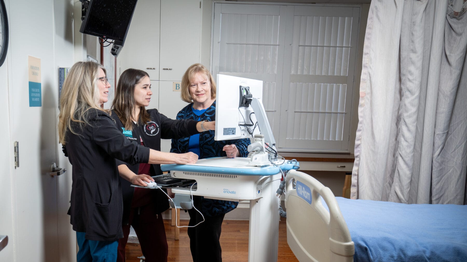 Three healthcare professionals stand around a computer on a mobile cart, discussing something on the screen, next to a hospital bed.