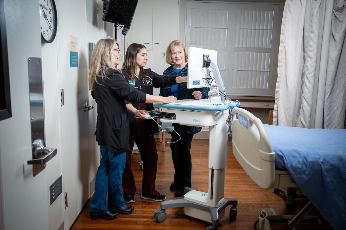 Three healthcare professionals interact with a medical monitor beside a hospital bed in a clinical setting.