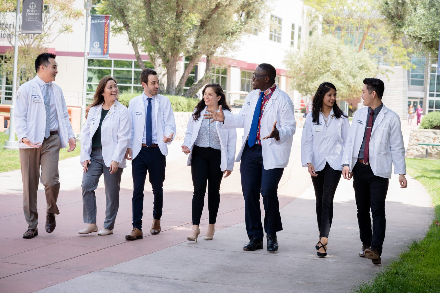 A group of medical professionals, wearing white lab coats, walk and converse in an outdoor setting near a medical facility.