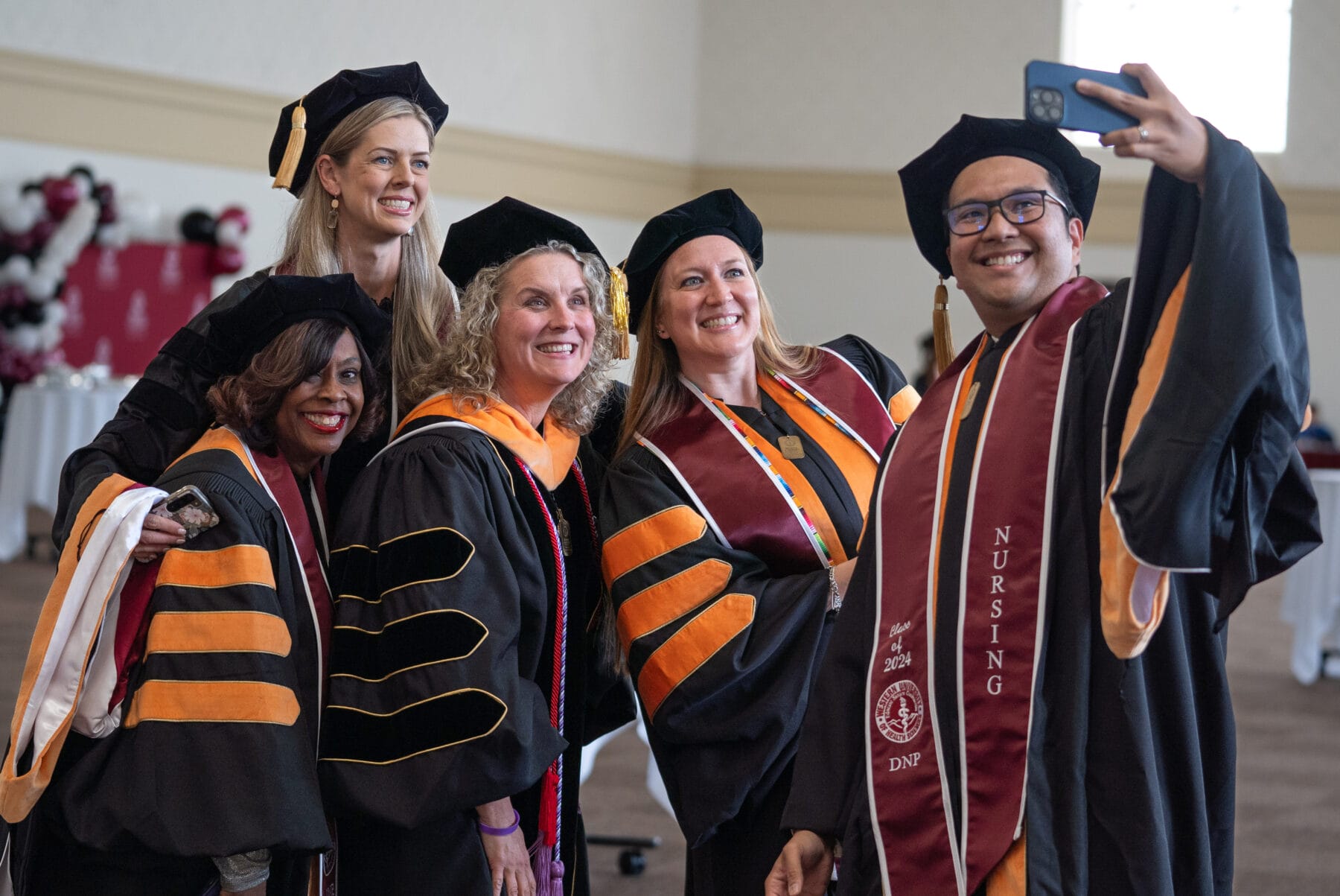 A group of people in academic regalia take a selfie together, celebrating a graduation event indoors.