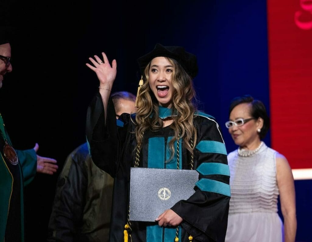 A graduate in ceremonial attire waves and holds a diploma, while standing on stage with a red backdrop and several other people.