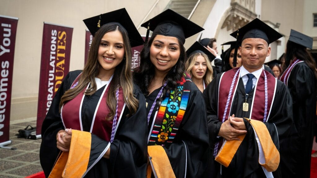 Graduates in caps and gowns smile while standing in line outside a building during a graduation ceremony.