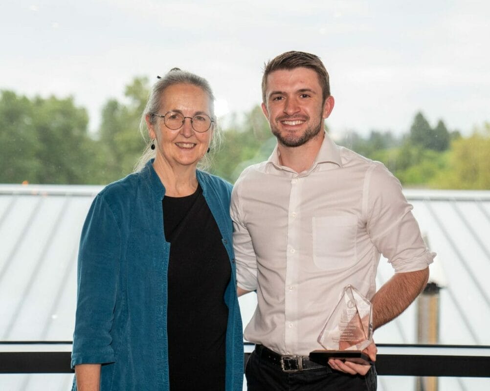 A woman in a blue jacket and a man in a white shirt stand side by side smiling. The man holds a clear glass award. They are indoors with large windows showing a tree and sky background.