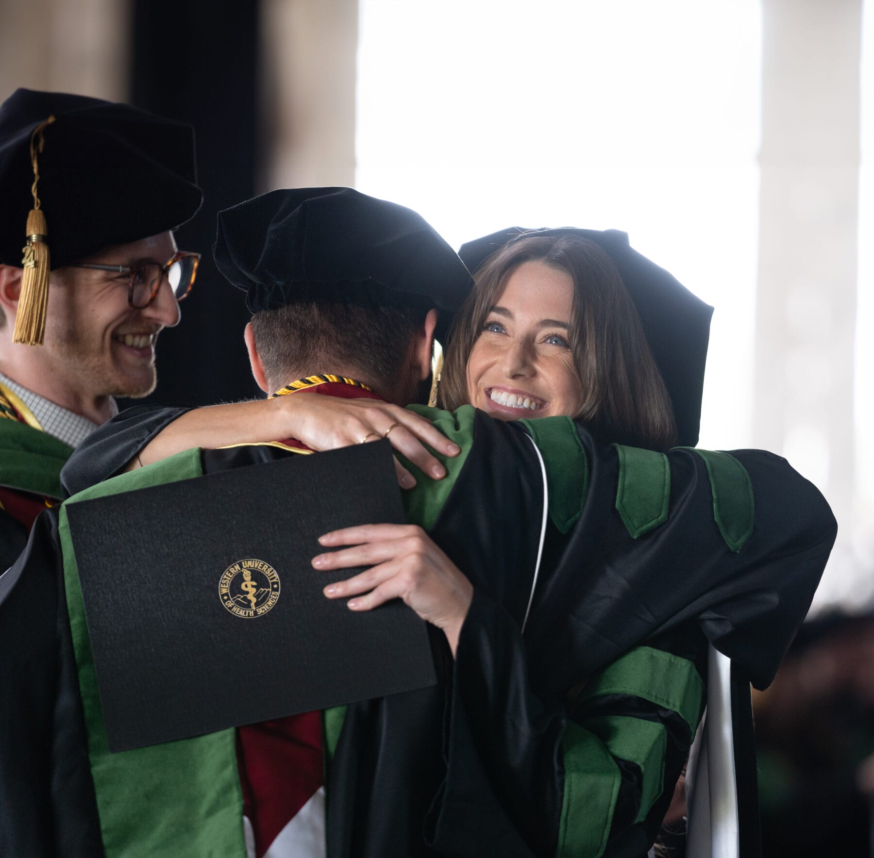 Two graduates in academic regalia embrace while holding a diploma, with another graduate smiling beside them at a graduation ceremony.