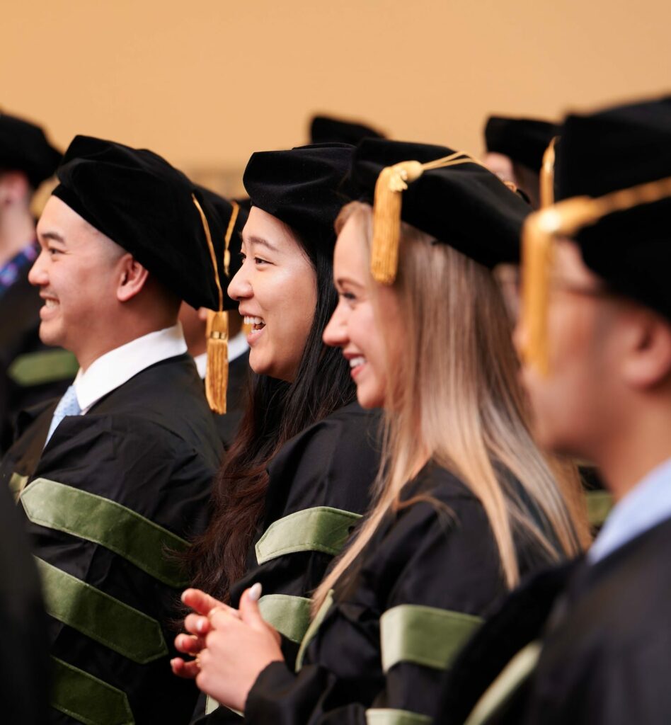 A group of graduates dressed in black robes with green accents and black caps with gold tassels sit and smile during a graduation ceremony.