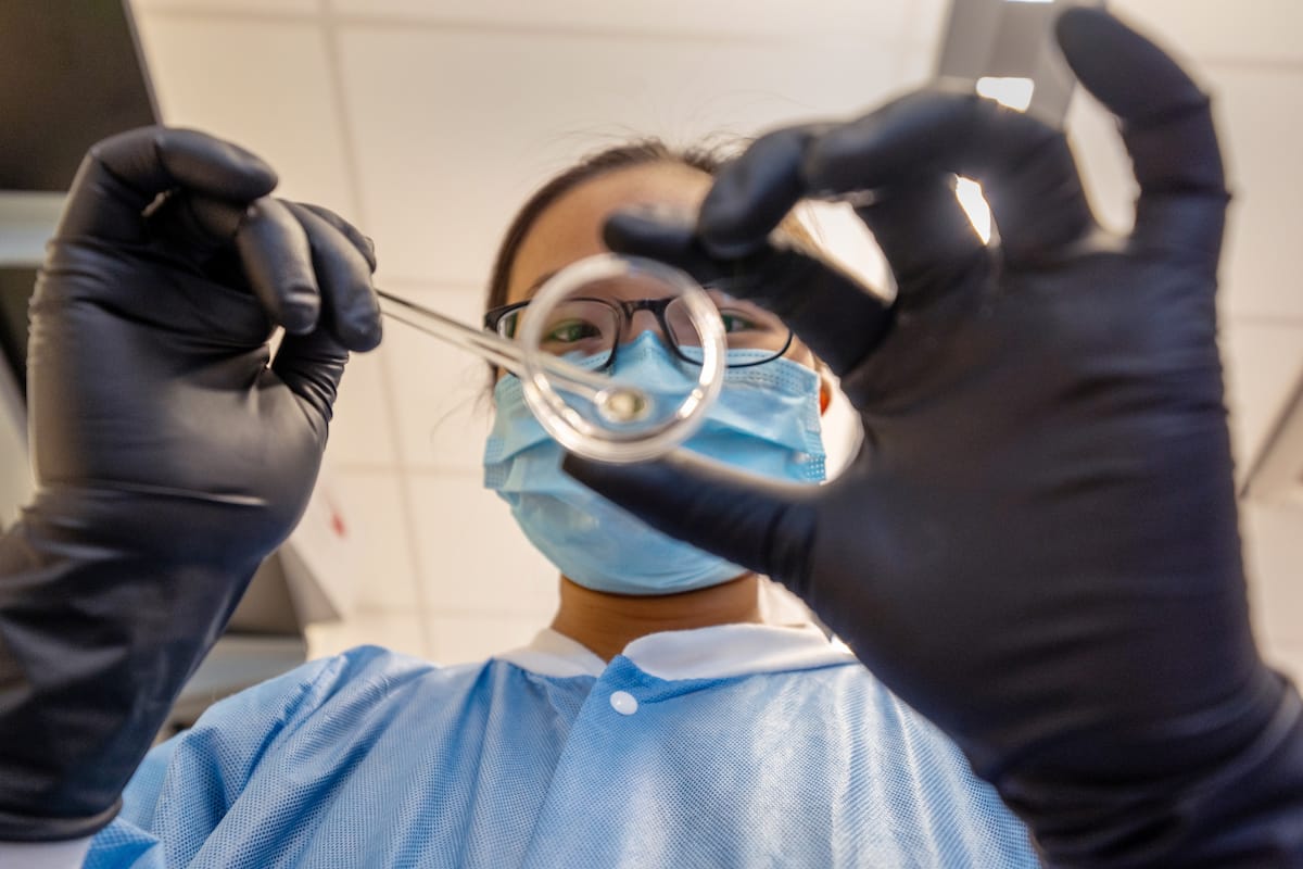 A person wearing a mask, protective gown, and black gloves holds up laboratory equipment, focusing on a small object in a lab setting.