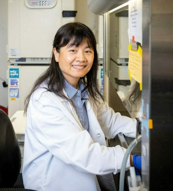 A woman wearing a lab coat sits at a workstation in a laboratory, smiling at the camera. Laboratory equipment and notes are visible in the background.