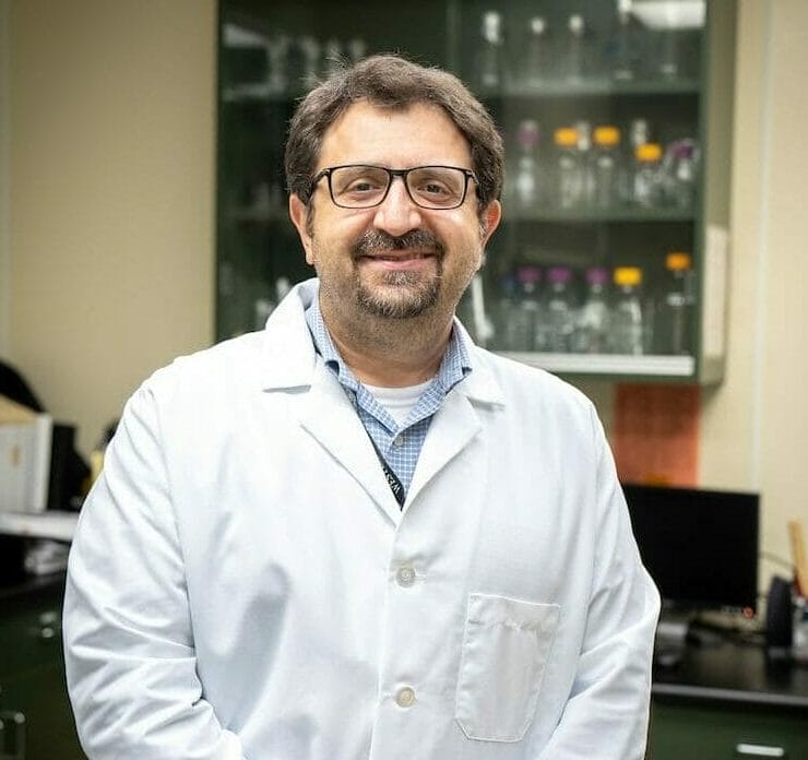 A person wearing glasses and a white lab coat stands in a laboratory with hands clasped. Shelves with bottles and equipment are visible in the background.