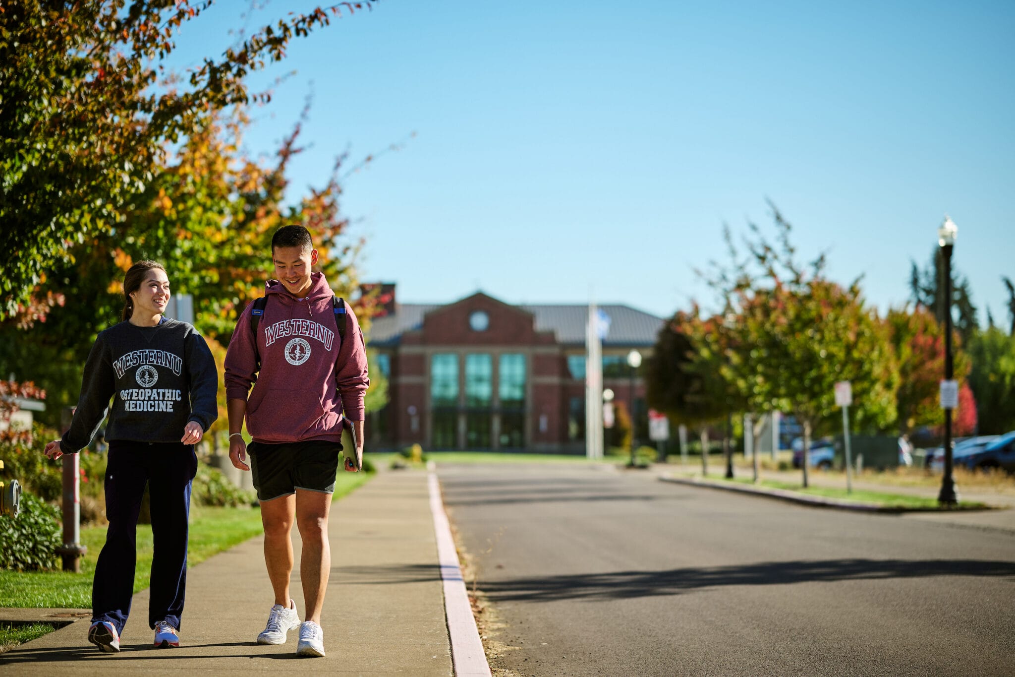 Two people wearing university sweatshirts walk and talk on a sunny campus pathway lined with trees, with a brick building visible in the background.