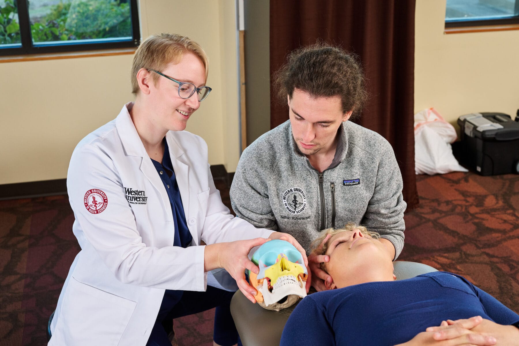 A person in a lab coat demonstrates with a model skull, while another person observes and a person lies on a table in a classroom setting.