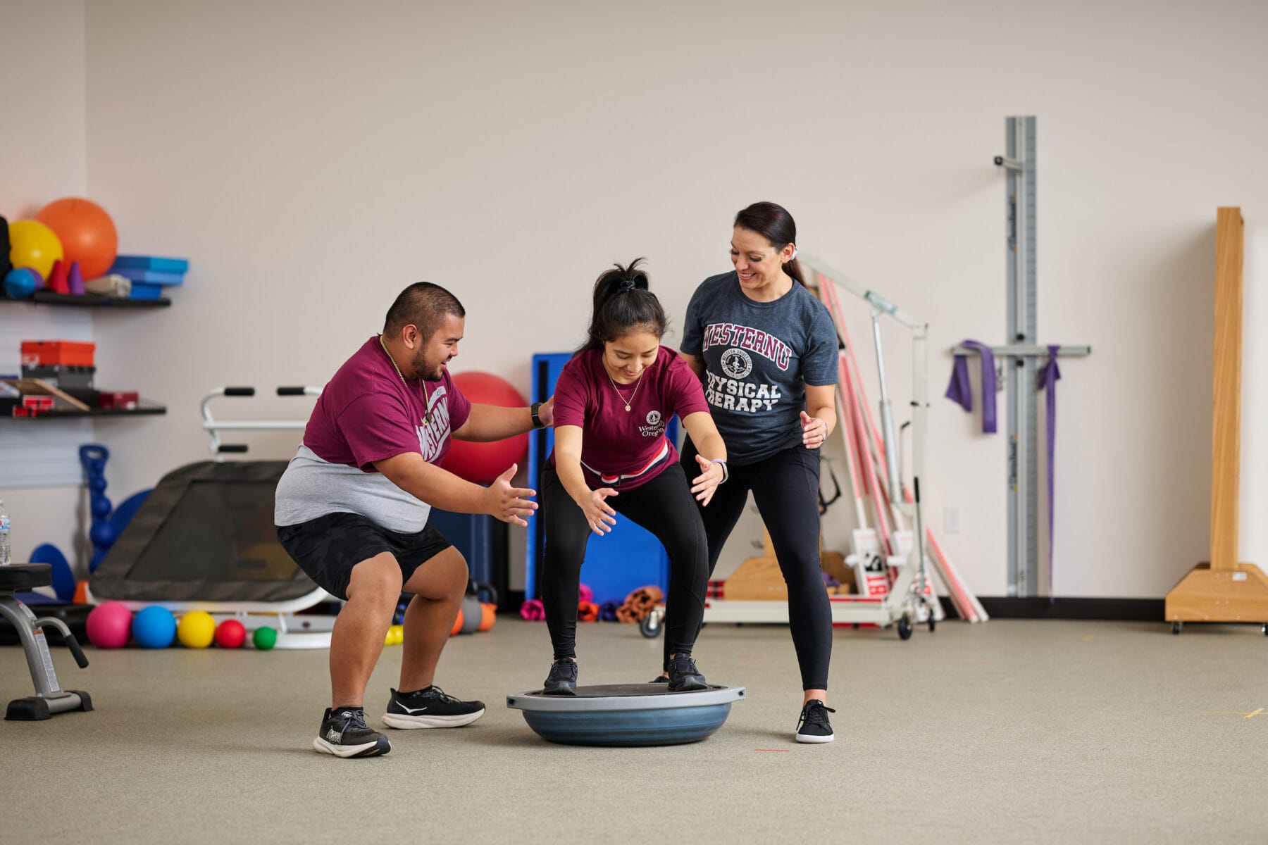 Two people assist a woman balancing on a Bosu ball in a physical therapy room filled with equipment.