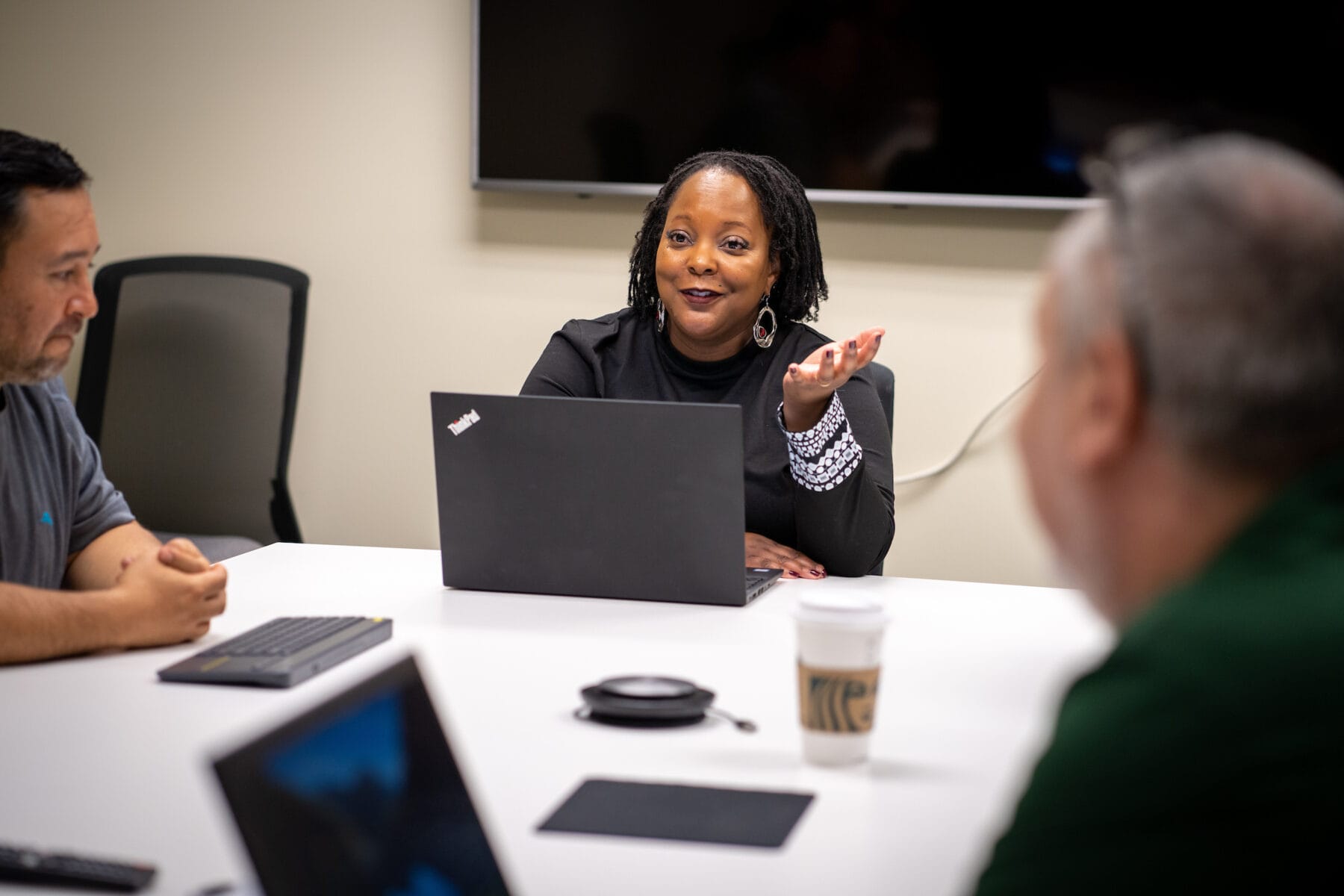 A woman sits at a conference table with a laptop, gesturing as she speaks to two colleagues during a meeting.
