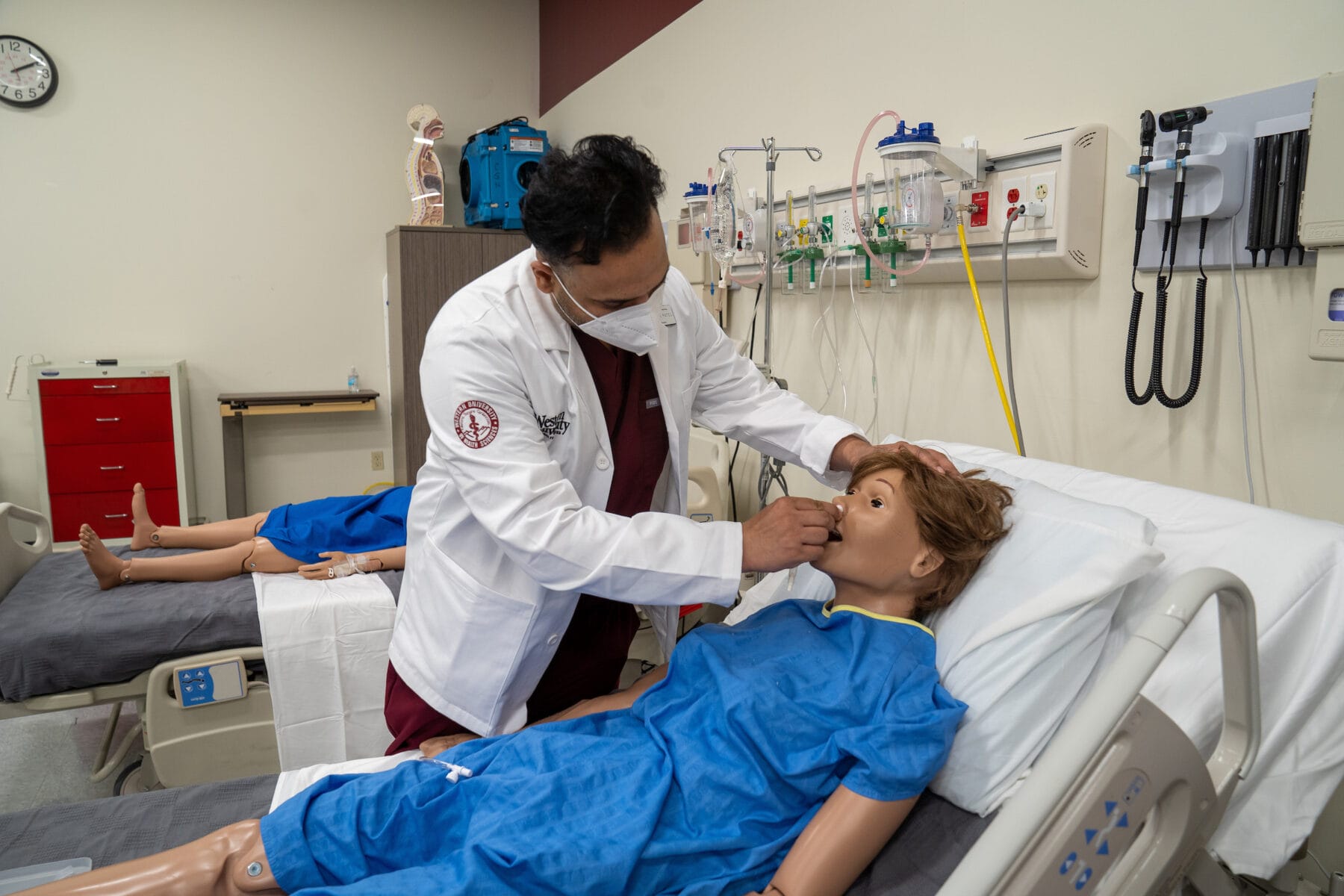 A medical professional practices a procedure on a medical mannequin in a simulation lab, with medical equipment and another mannequin in the background.