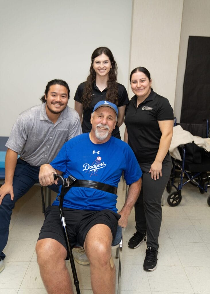 A man with a cane sits and smiles, surrounded by three standing people. One wears a Dodger's shirt and hat. They are indoors.