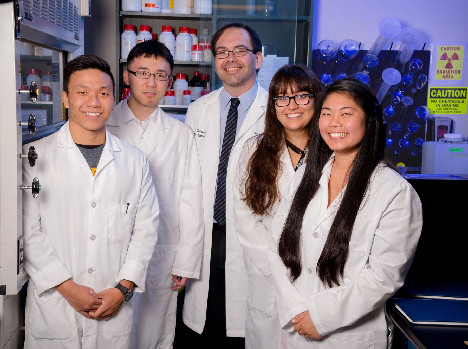 A group of five people in lab coats stand together in a laboratory, smiling at the camera. Shelves with bottles and lab equipment are visible in the background.